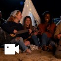 Sześć osób siedzących na plaży i jedna dziewczyna trzyma gitarę